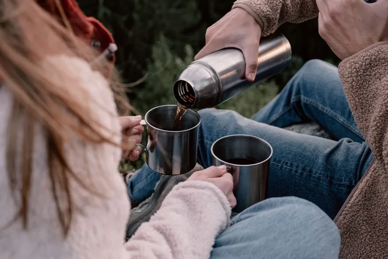 Sirviendo bebida caliente desde un termo de acero inoxidable en tazas metálicas al aire libre